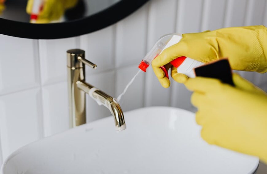 From above anonymous housekeeper in yellow rubber gloves spraying cleaner to chrome water faucet in bathroom